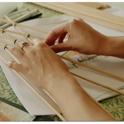 Hands tying thin bamboo rods together into a scale model on a drawing table