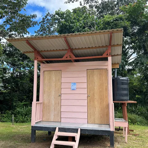 A pink-painted public bathroom raised on stilts with two wooden doors, steps up to the platform and a water tank feeding a washing point at the side