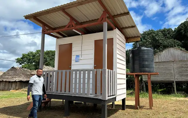 Een afgebouwd openbaar toiletgebouw op palen bij de krutu oso, met golfplaten dak, twee deuren en een zwarte watertank op een houten stellage ernaast