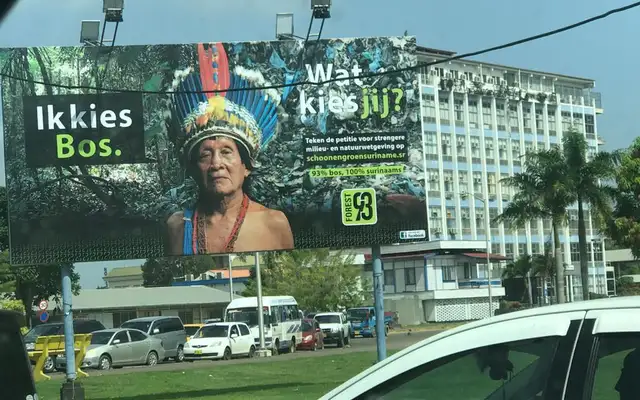 A roadside billboard on the Sophie Redmondstraat: an indigenous elder in a feather headdress, half against forest and half against a wall of plastic waste, headed "Ik kies Bos." and "Wat kies jij?" with a call to sign the petition at schoonengroensuriname.sr