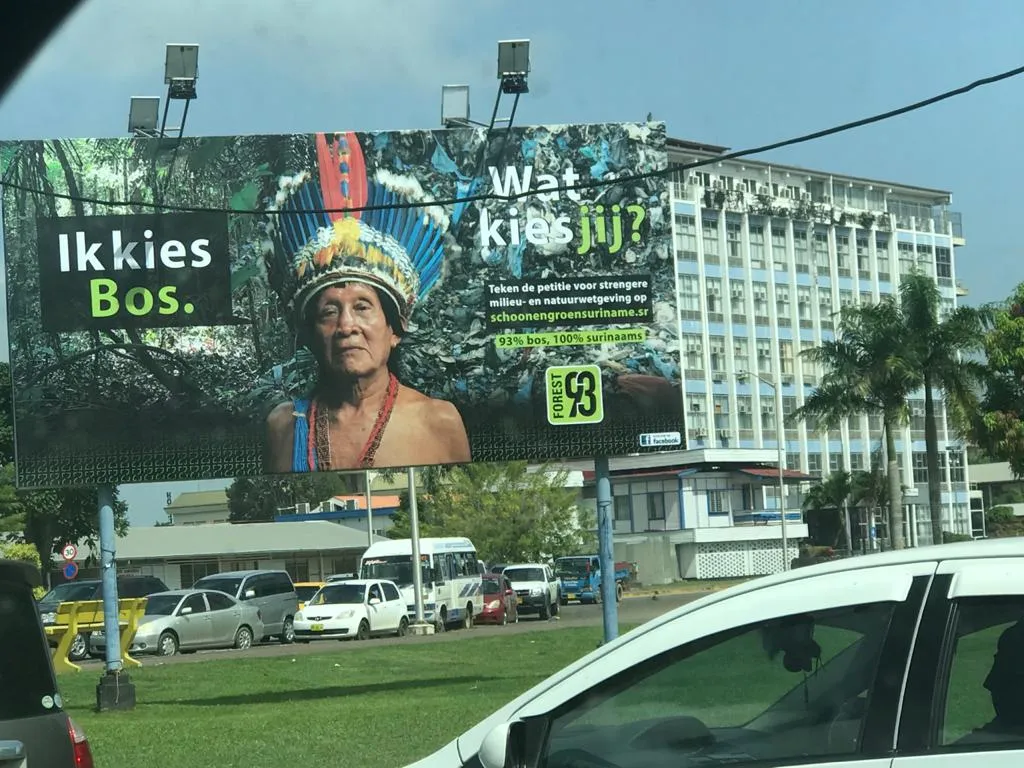 A roadside billboard on the Sophie Redmondstraat: an indigenous elder in a feather headdress, half against forest and half against a wall of plastic waste, headed "Ik kies Bos." and "Wat kies jij?" with a call to sign the petition at schoonengroensuriname.sr