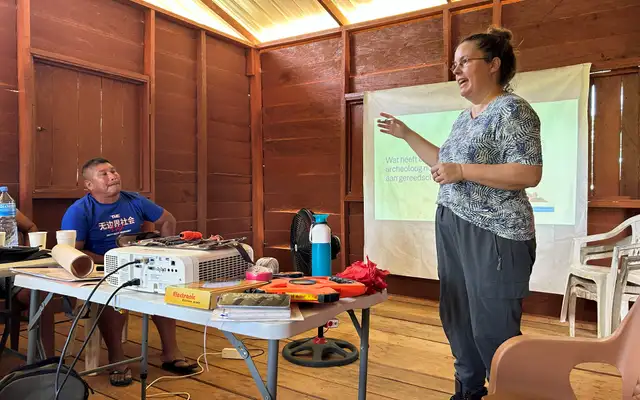 Irene Meulenberg geeft de training gemeenschapsarcheologie in een houten zaal, naast een tafel met meetgereedschap en een dia met de vraag welk gereedschap een archeoloog nodig heeft