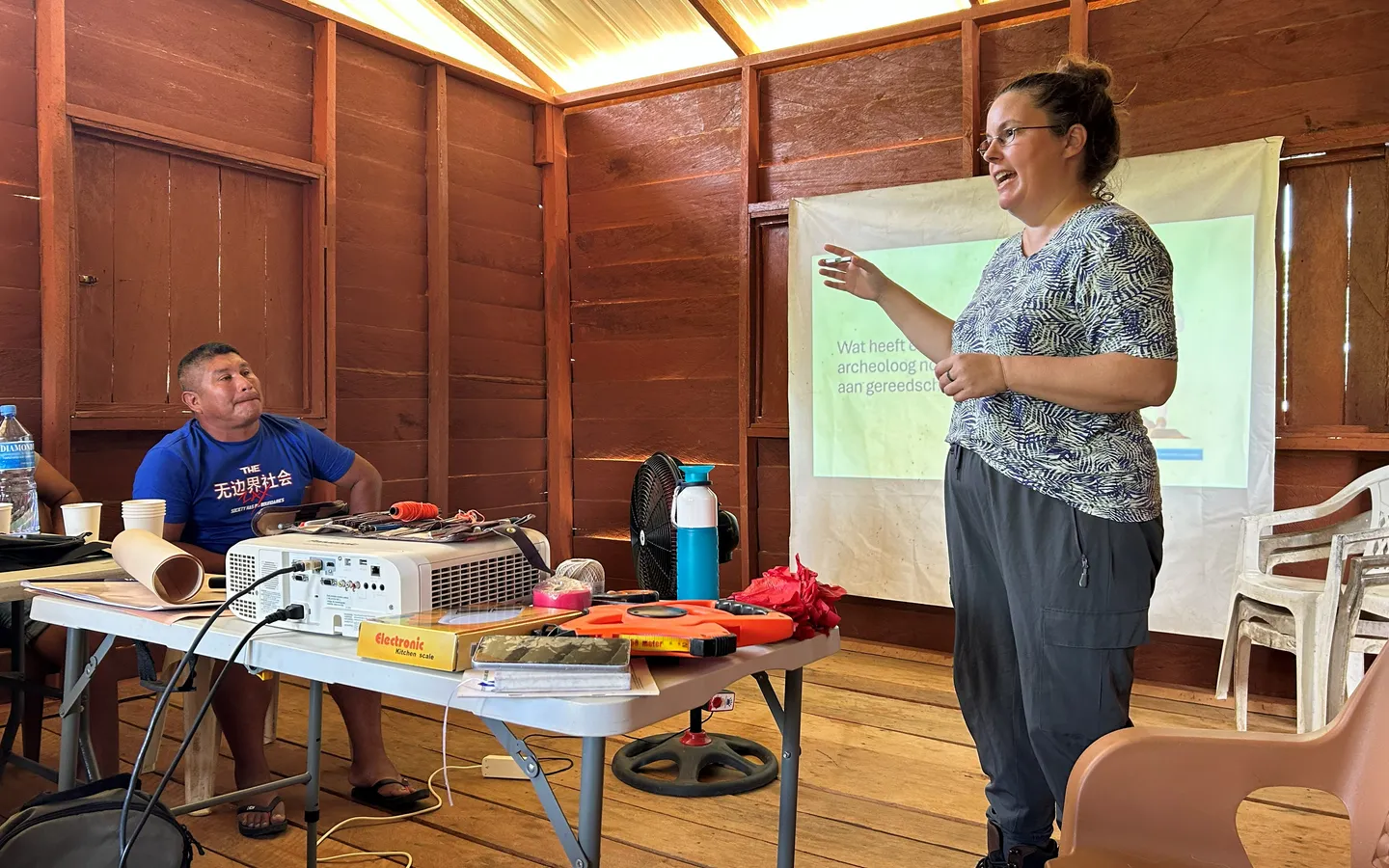 Irene Meulenberg teaching the community archaeology training in a wooden hall, beside a table of survey tools and a projected slide asking what tools an archaeologist needs