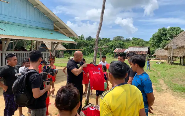 Dean Gorré holding up a red football shirt for a group of young people gathered around him in the village, with a camera operator filming beside him
