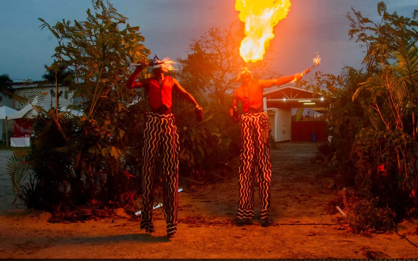 Two stilt walkers in red and striped costumes breathing fire at dusk at the Urban Heritage & Art Festival, under a 93% Bos. 100% Surinaams. banner