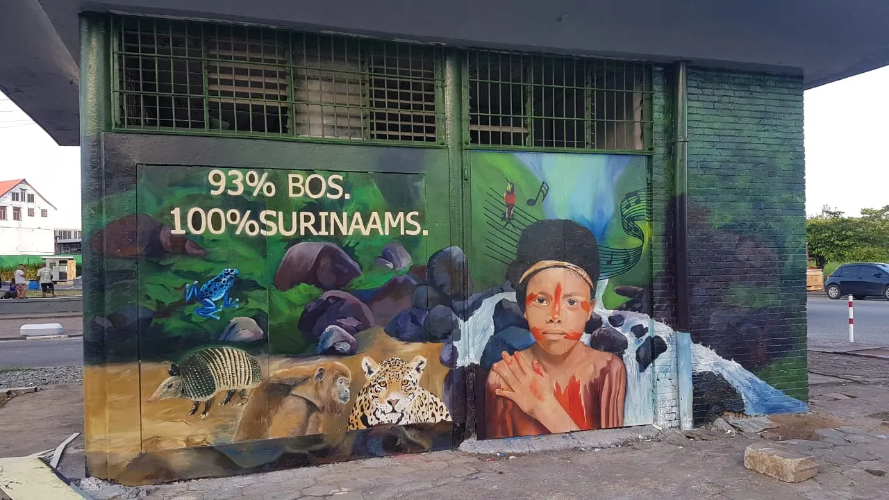 The finished mural on the utility building: "93% BOS. 100% SURINAAMS." painted above a blue poison dart frog, an armadillo, a monkey and a jaguar, beside a young girl in red face paint with a waterfall behind her