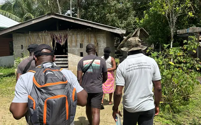Deelnemers aan het oriëntatiebezoek lopen door Tottikamp naar een huis van betonblokken, een van hen in een shirt van de Dienst Watervoorziening van het Ministerie van Natuurlijke Hulpbronnen