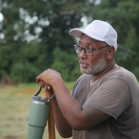 A man in a white cap leans on a spade at the planting site