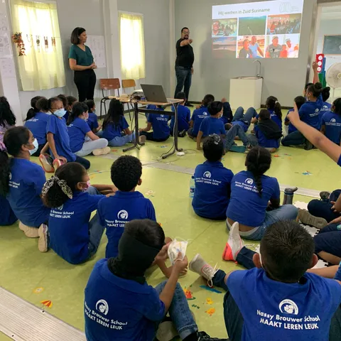 Leerlingen van de Nassy Brouwer School in Paramaribo zitten op de grond tijdens een les over inheemsen in Suriname, bij een dia met de kop “Wij werken in Zuid Suriname”