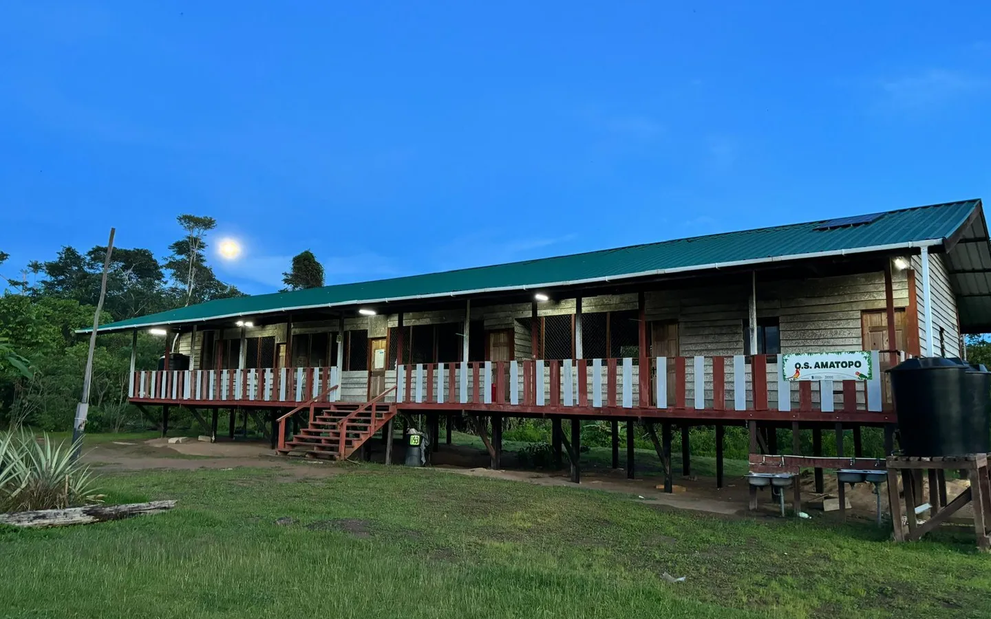 The school at Amatopo at dusk with its veranda lights on, the moon above the treeline behind it
