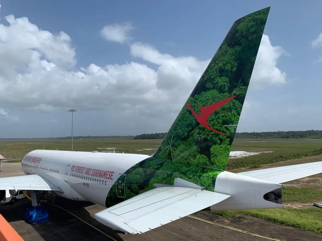 The tail of the Surinam Airways Boeing 777 on the apron, printed with an aerial view of dense green forest behind the red bird logo, and "93% FOREST 100% SURINAMESE." along the fuselage