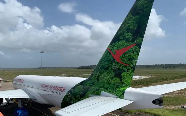 The tail of the Surinam Airways Boeing 777 on the apron, printed with an aerial view of dense green forest behind the red bird logo, and "93% FOREST 100% SURINAMESE." along the fuselage