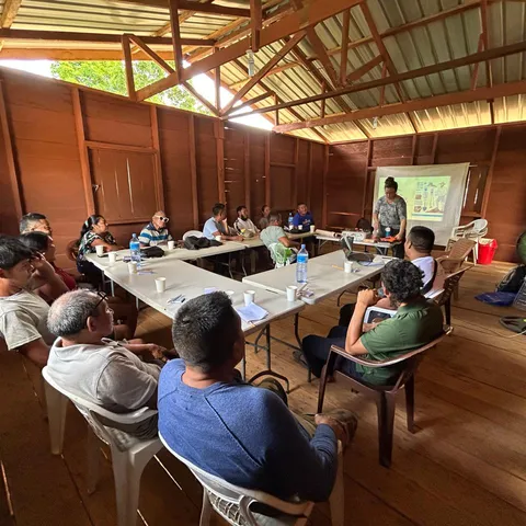 A meeting under way inside the Tewe office in August 2026, villagers seated around tables in the timber hall while a presenter speaks at a screen